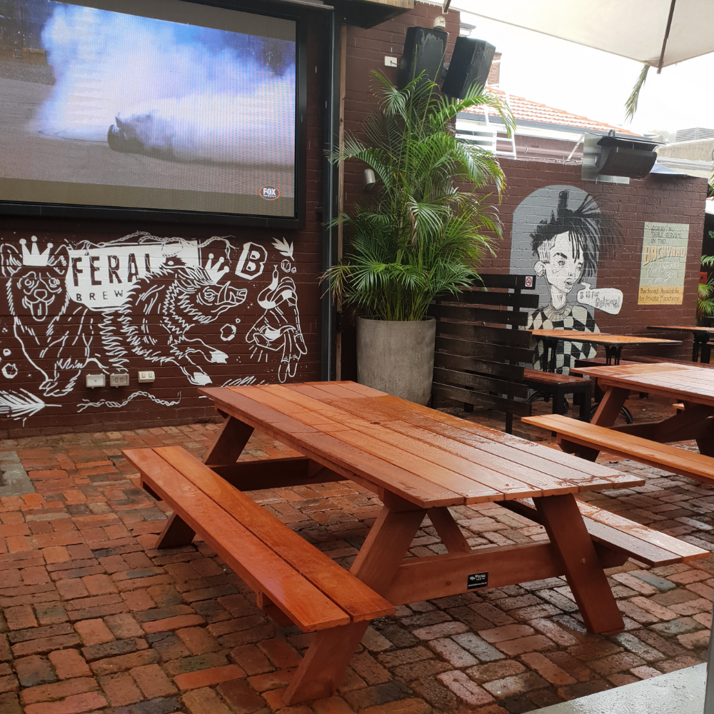 Outdoor seating area with hardwood picnic tables and a large screen displaying content, featuring a mural of a bear and a person.