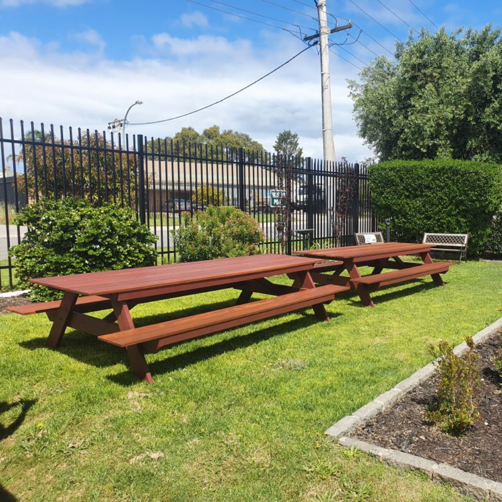 Two 12 seater A frame Jarrah picnic tables on a grassy area with a fence and trees in the background.