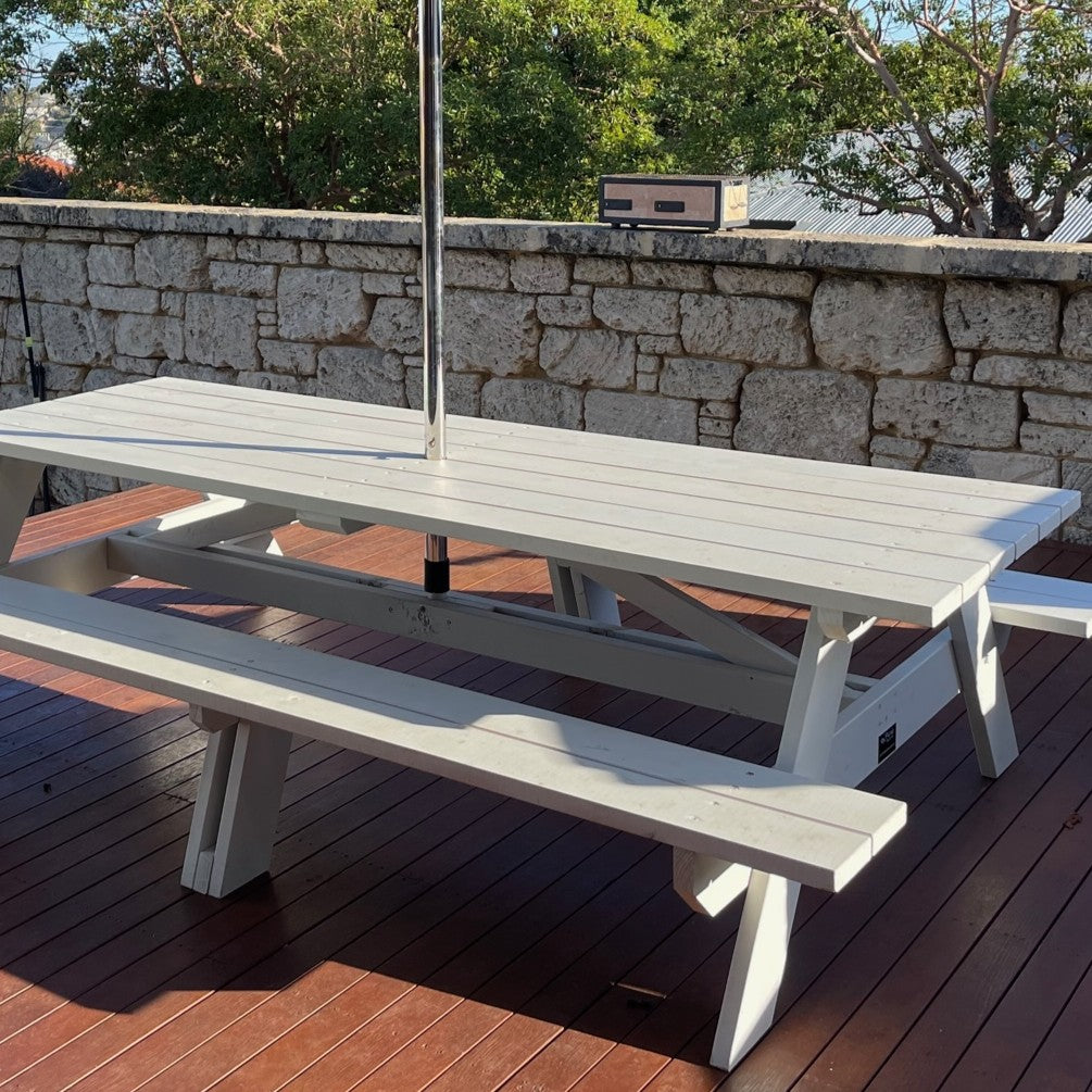 A white outdoor picnic table with attached benches on a wooden deck, with a stone wall, trees, and a grill in the background.