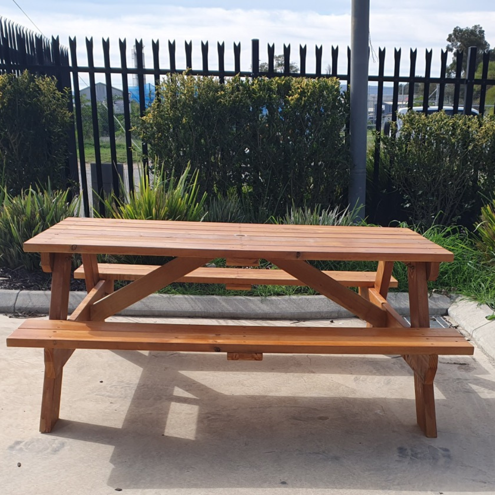 Wooden picnic table with benches on a concrete surface, surrounded by greenery and a fence.