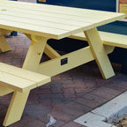 A light yellow wooden picnic table with attached benches, photographed outdoors on a paved surface with a blue wall in the background.