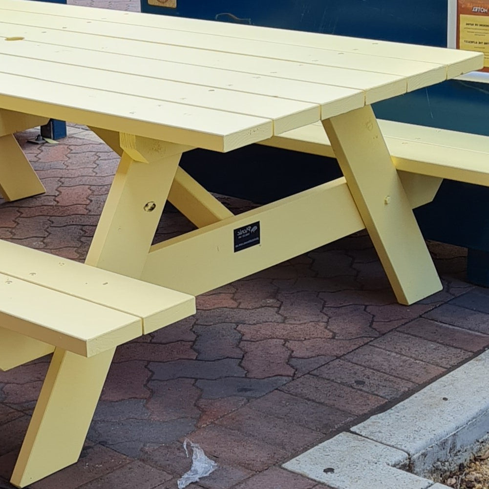 A light yellow wooden picnic table with attached benches, photographed outdoors on a paved surface with a blue wall in the background.
