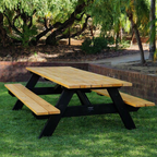 A wooden picnic table with attached benches, featuring light natural wood tops and painted black A-frame legs, situated on a green lawn in an outdoor backyard setting with trees and a brick retaining wall.
