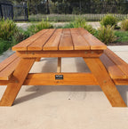 A wooden picnic table with attached benches, shown outdoors on a concrete surface with plants and a black metal fence in the background. A small black brand plaque is visible on the front leg.